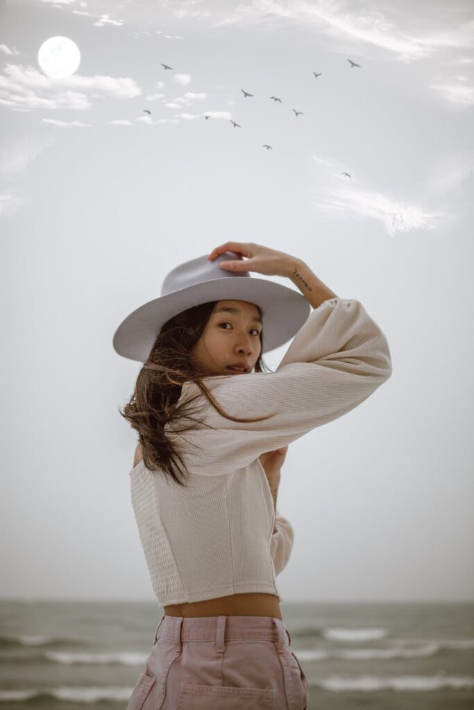 peaceful young asian lady adjusting hat near stormy ocean